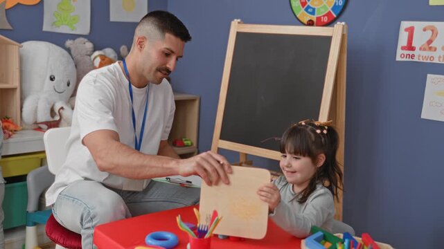 Man teacher hands toy to girl at preschool table in classroom building while taking notes on a clipboard and guiding play; gentle guidance calm.