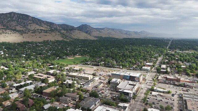 Aerial view of the city of Boulder nestled against the Flatirons with lush green trees, urban buildings, and mountain ranges under a cloudy sky in Boulder, Colorado, United States.