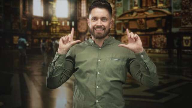 Hispanic man frames his face with hands and smiles inside an ornate museum hall, making l shaped gestures toward camera; playful curiosity.