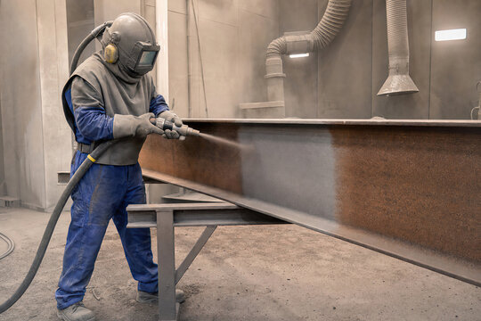 Sandblaster cleans rusty steel beam with abrasive tool. Protective mask and gear worn by worker in factory. Metal surface preparation, industrial maintenance and corrosion removal.