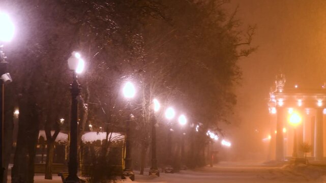 Fabulous view of the snow-covered city park at night, with trees in the snow, cozy warm light of lanterns.