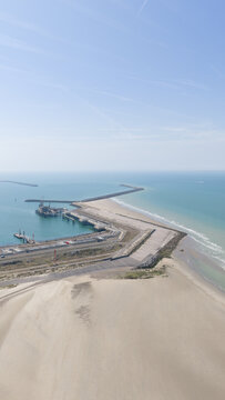 Aerial view of Dunkirk LNG Terminal featuring industrial infrastructure, storage tanks, and jetties extending into the North Sea, with sandy coastline. Loon-Plage, Hauts-de-France, France.