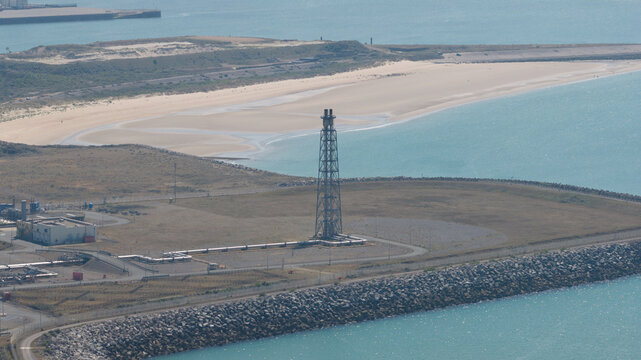 Aerial view of the Dunkirk LNG Terminal featuring its prominent flare stack, industrial infrastructure, and coastal setting near the sandy beach and sea in Loon-Plage, Hauts-de-France, France.