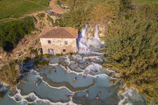 Aerial view of Cascate del Mulino with people bathing in the tiered thermal pools next to an old stone mill building in Terme di Saturnia, Tuscany, Italy.