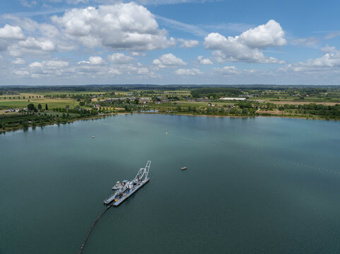 Aerial view of a dredging vessel operating on a calm lake surrounded by lush green fields and trees under a bright blue sky with scattered white clouds in the Netherlands.