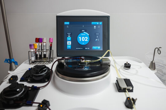 The touchscreen of an apheresis machine shows the process data beside a rack of blood tubes during a plasma donation at a clinic.