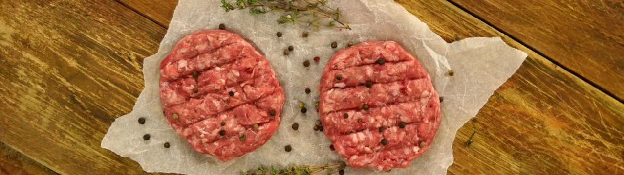 Two uncooked beef patties seasoned with peppercorns and thyme resting on parchment paper over a rustic wooden table.