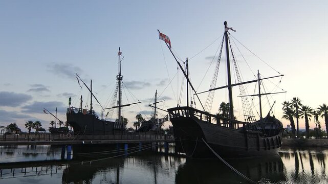 Cinematic wide shot of the historical replica ships. Pinta, Ni&ntilde;a and Santa Mar&iacute;a docked at Muelle de las Carabelas during a serene sunset. Located in La Rabida, Palos de la Frontera, Huelva, Spain.