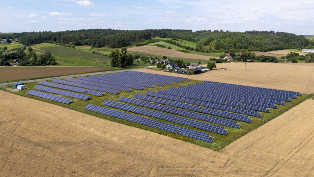 Aerial view of a solar farm with rows of photovoltaic panels situated in a rural landscape surrounded by golden wheat fields and green trees in Grudziadz, Kuyavian-Pomeranian, Poland.
