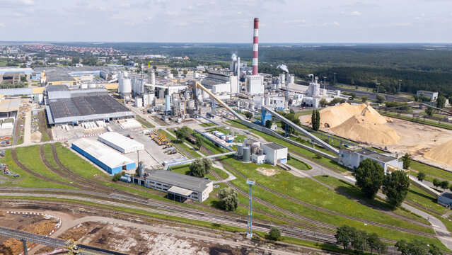 Aerial view of a large industrial paper mill facility featuring a tall chimney, extensive storage piles of wood chips, and surrounding rail infrastructure in Grudziadz, Kuyavian-Pomeranian, Poland.