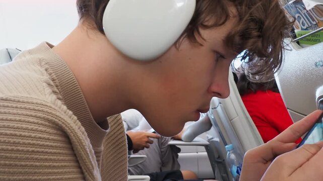 A young man with headphones leans forward while using a smartphone on an airplane, nearby passengers seated in the rows ahead, with a water bottle on the tray table