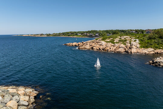 Aerial view of a small sailboat navigating the calm blue waters of Rockport Harbor, framed by rugged rocky coastlines and lush green vegetation in Rockport, Massachusetts, United States.