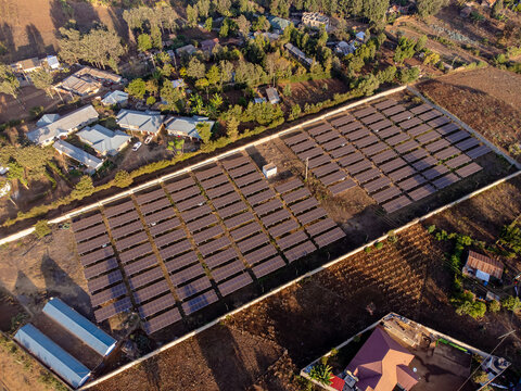 Aerial view of a solar farm with rows of photovoltaic panels in a fenced enclosure near residential buildings in Loitokitok, Kajiado County, Kenya.