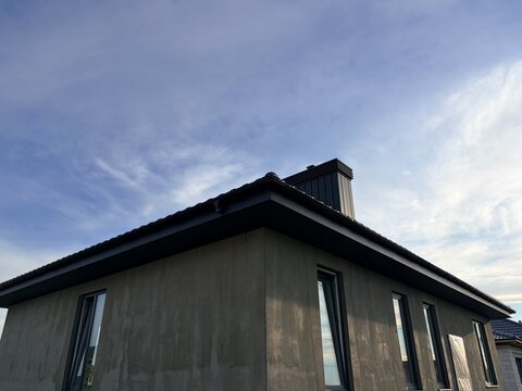 A house with grey plaster walls and a black metal roof against a blue sky. House construction