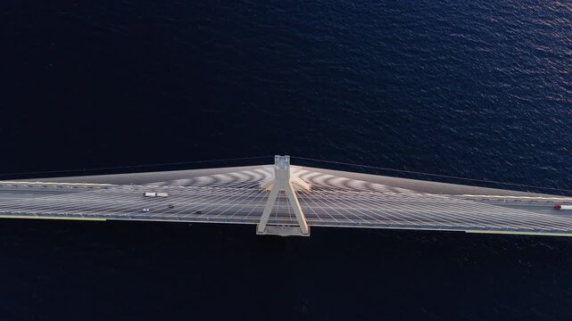 Top details of Rio-Antirrio Bridge crossing Gulf of Patras at sunset, drone tilt backward reveals beams on sparkling deep blue water
