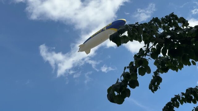 A Goodyear blimp flies across a blue sky with white clouds, framed by green tree leaves in London, England