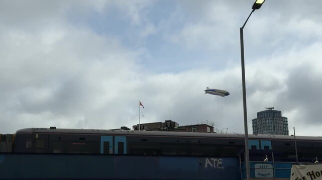 A Goodyear blimp flies over a passing train in London, England, on a cloudy day