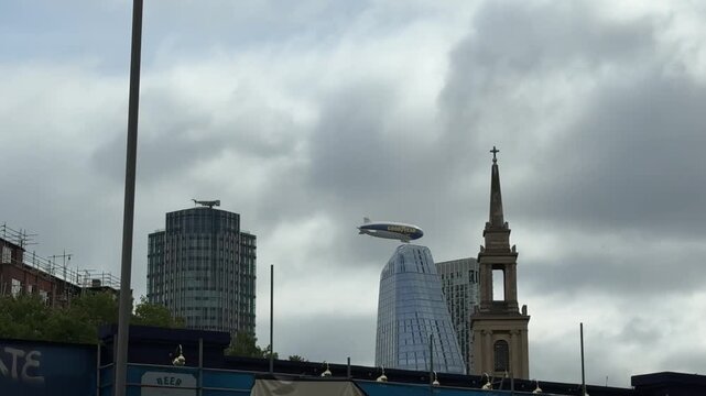 The Goodyear blimp flies across the London skyline with modern skyscrapers and a church steeple