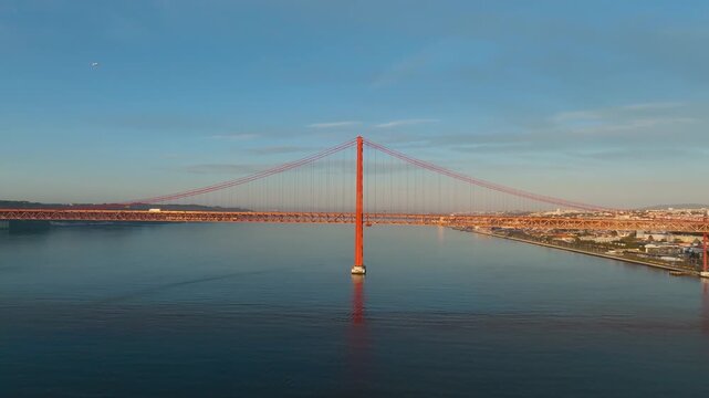 Panoramic drone pan shot of 25 de Abril Bridge, Lisbon. Golden sunrise light on Tagus River. Clear day. Portugal