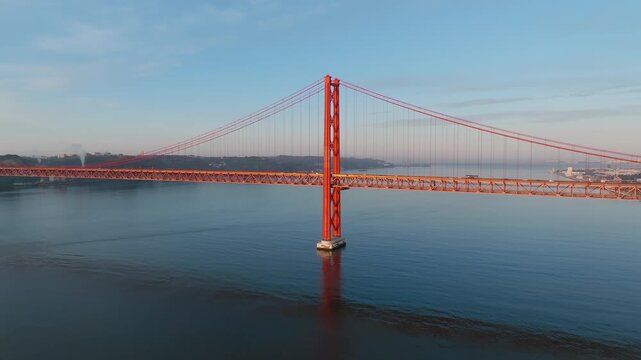 25 de Abril Bridge, Lisbon. Panoramic drone pan at sunrise. Warm golden light, Tagus River below, clear sky. Portugal