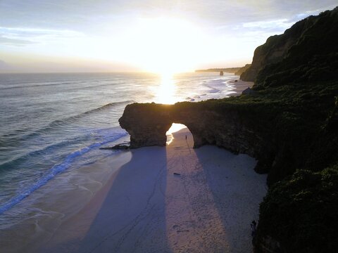 Aerial view of a natural rock arch on a sandy beach at sunset with calm waves and lush cliffs in Indonesia.