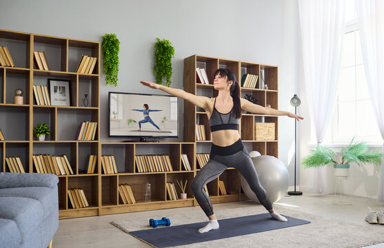 Woman yoga home fitness exercise training balance. She performs a warrior pose on a mat, using a tv workout, building strength, flexibility, stretching and posture. Healthy life at home concept.