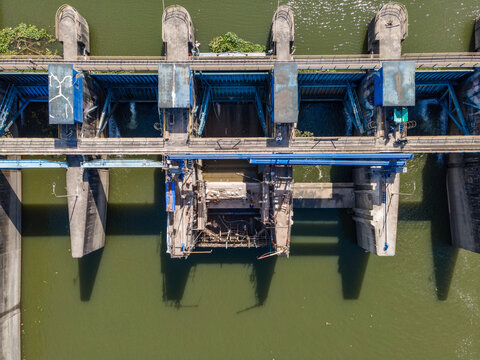 Aerial view of a concrete dam with blue metal sluice gates and flowing river water in Banyumas Regency, Central Java, Indonesia.
