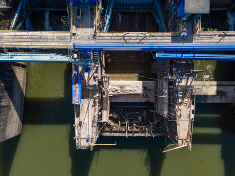 Aerial view of a dam construction site with metal scaffolding and a blue gantry crane over the water in Banyumas Regency, Central Java, Indonesia.