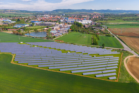Aerial view of a solar farm with rows of photovoltaic panels in green fields near industrial buildings and rolling hills in Kyjov, South Moravian Region, Czechia.