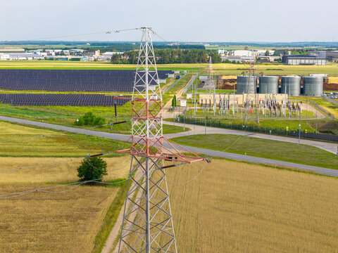 Aerial view of a high-voltage electricity pylon standing amidst agricultural fields, with a solar panel farm and an electrical substation in the background in Poland.