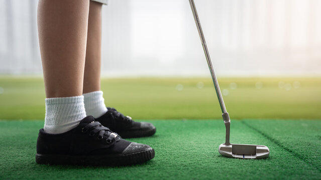 Junior Golfer standing with a putter on a green practice mat at a driving range in a close-up view of an Asian child's legs, shoes, and golf equipment