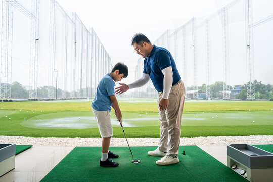 Junior Golf Lesson with a male coach giving posture guidance to an Asian boy holding a club on a practice mat at a spacious outdoor driving range
