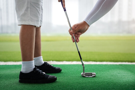 Golf Coach correcting an Asian boy's putter position and hand placement during a close-up junior golf lesson on artificial turf at a driving range