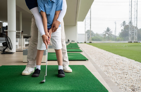 Junior Golf Lesson with a male coach standing behind an Asian boy to correct his grip and club alignment on a practice mat at a covered driving range