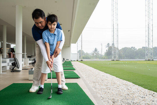 Junior Golf Lesson with a male coach standing behind an Asian boy to guide his hands, grip, and club alignment during practice at a covered driving range