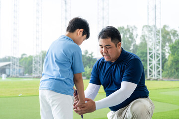 Fototapeta premium Junior Golf Lesson with a male golf coach kneeling to correct an Asian boy's grip and club position during practice at a bright outdoor driving range