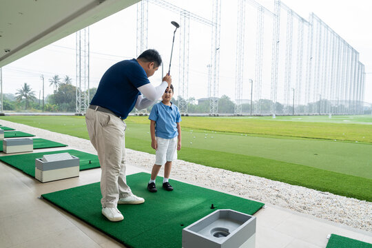 Golf Coach demonstrating swing technique to an Asian boy who watches attentively during a junior golf lesson on a practice mat at a driving range