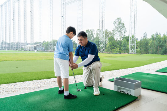 Junior Golf Lesson with a male golf coach kneeling beside an Asian boy to adjust his club position and stance on a practice mat at a driving range