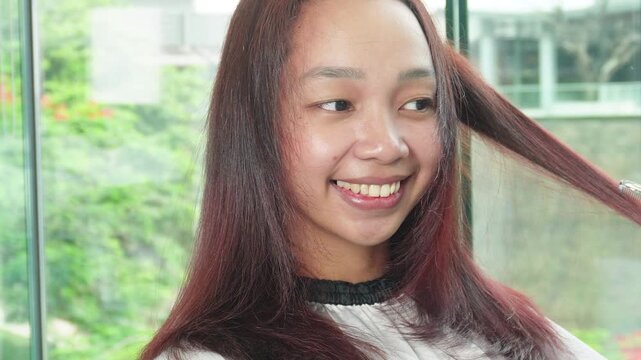 Hair stylist trimming and styling reddish brown hair after professional hair coloring treatment for Southeast Asian woman in her early 20s