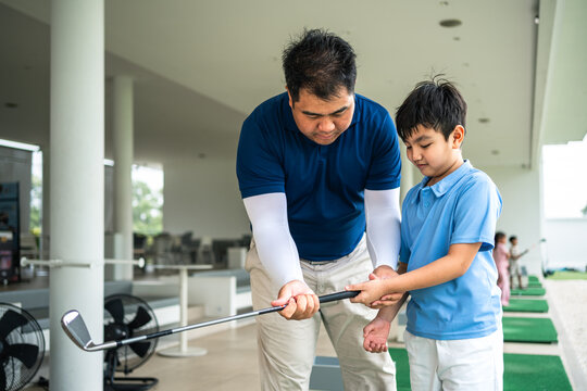 Golf Coach teaching an Asian boy the correct club grip through hands-on guidance during a junior golf lesson at a bright covered driving range
