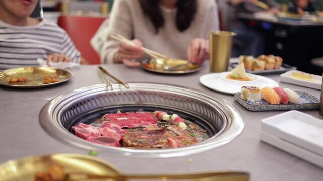Family enjoying korean bbq dinner with beef, sushi, and nigiri in a japanese restaurant while grilling meat on a tabletop stove and using chopsticks for eating