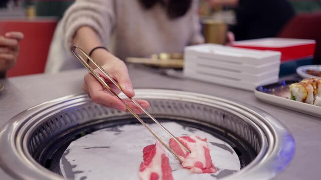Woman grilling delicious raw meat slices on a tabletop grill at an asian barbecue restaurant, enjoying a gourmet dining experience with traditional food preparation