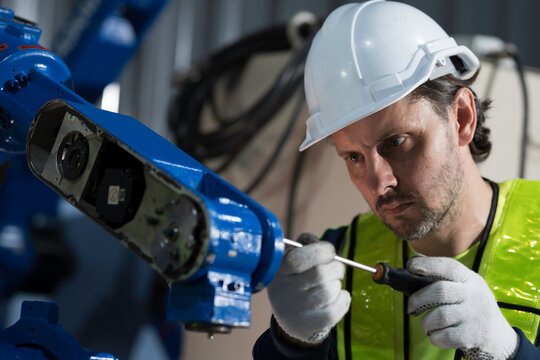 Male engineer wearing uniform, white hard hat and gloves repair automatic robotic arm for welding. Male technician inspecting quality automatic welding robot arm machine with hand tools