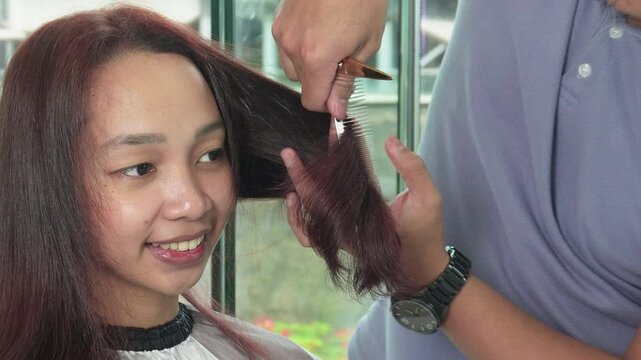 Hair stylist trimming and styling reddish brown hair after professional hair coloring treatment for Southeast Asian woman in her early 20s