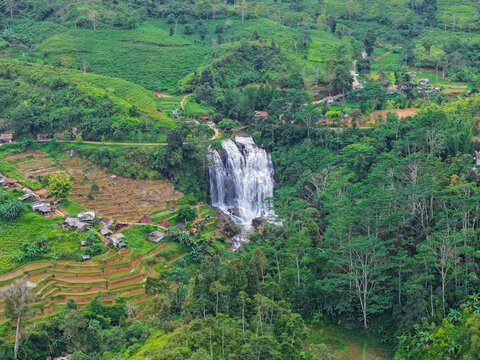 Aerial view of Curug Cikondang waterfall surrounded by lush green rice terraces, tropical forests, and small rural houses in the hills of Campaka, West Java, Indonesia.