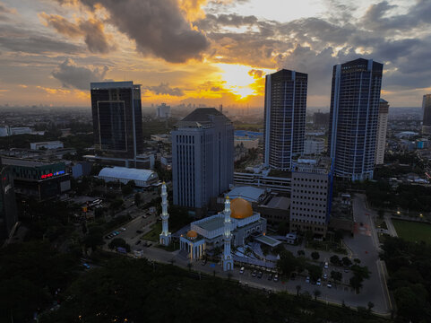 Aerial view of Jami' Assahara Mosque surrounded by modern skyscrapers and urban infrastructure during a vibrant sunset in Kecamatan Kembangan, Daerah Khusus Ibukota, Indonesia.