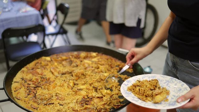 Woman serving traditional spanish paella from a large paellera pan onto a plate during a family meal. Close-up view of hands putting the rice dish on a ceramic plate