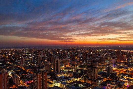 Aerial view of the Asuncion cityscape at sunset, featuring illuminated buildings, high-rise architecture, and the glowing horizon over the Paraguay River. Asuncion, Asuncion, Paraguay.