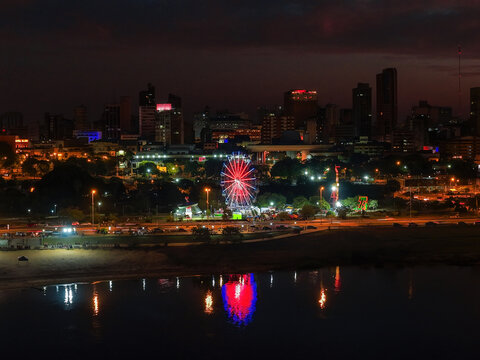 Aerial view of the illuminated ferris wheel at EcoPark reflecting on the water at night with the city skyline in the background in Asuncion, Asuncion, Paraguay.