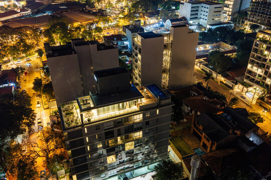 Aerial view of a modern residential building in Las Lomas at night, with illuminated terraces and rooftop pool, surrounded by city lights and trees in Asuncion, Asuncion, Paraguay.